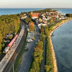 Pociąg Polregio Gdynia Hel stoi na stacji, otoczony lasem i morzem. W tle widać nadmorską miejscowość.