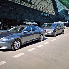 Dwa samochody, sedan i van, czekają przed nowoczesnym budynkiem lotniska Chopina w Warszawie. Może to być transport z Torunia.