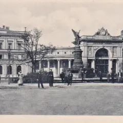 Dawny dworzec Wrocław Świebodzki z fontanną i pomnikiem. Ludzie w strojach z epoki spacerują przed imponującą budowlą.