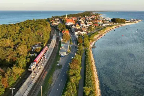 Pociąg Polregio Gdynia Hel stoi na stacji, otoczony lasem i morzem. W tle widać nadmorską miejscowość.