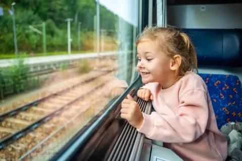Uśmiechnięta dziewczynka patrzy przez okno pociągu. Zastanawia się, od ilu lat można jeździć samemu pociągiem, marząc o podróżach.