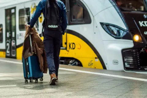 Podróżny z walizką i kurtką idzie w stronę nowoczesnego pociągu. Artus pociąg czeka na pasażerów.