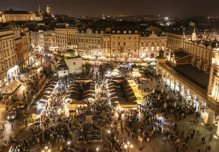 Tłumy ludzi spacerują po rozświetlonym jarmarku bożonarodzeniowym krakow. Wokół stragany, zabytkowe kamienice i Sukiennice.