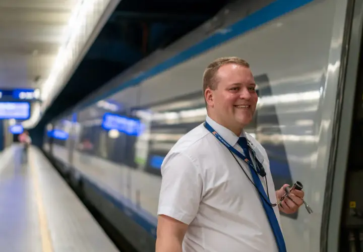 Uśmiechnięty kierownik pociągu w białej koszuli i granatowym krawacie stoi na peronie obok pociągu PKP Intercity.