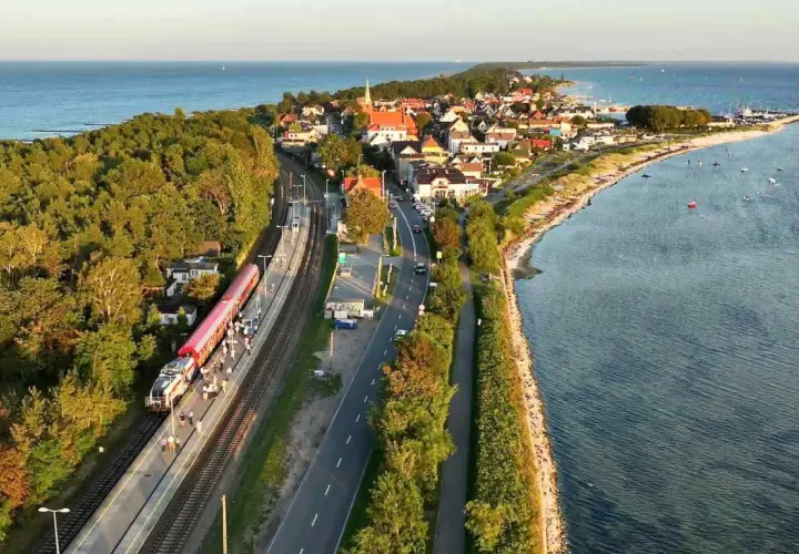 Pociąg Polregio Gdynia Hel stoi na stacji, otoczony lasem i morzem. W tle widać nadmorską miejscowość.