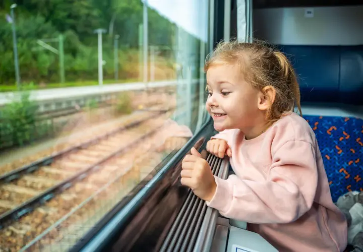 Uśmiechnięta dziewczynka patrzy przez okno pociągu. Zastanawia się, od ilu lat można jeździć samemu pociągiem, marząc o podróżach.
