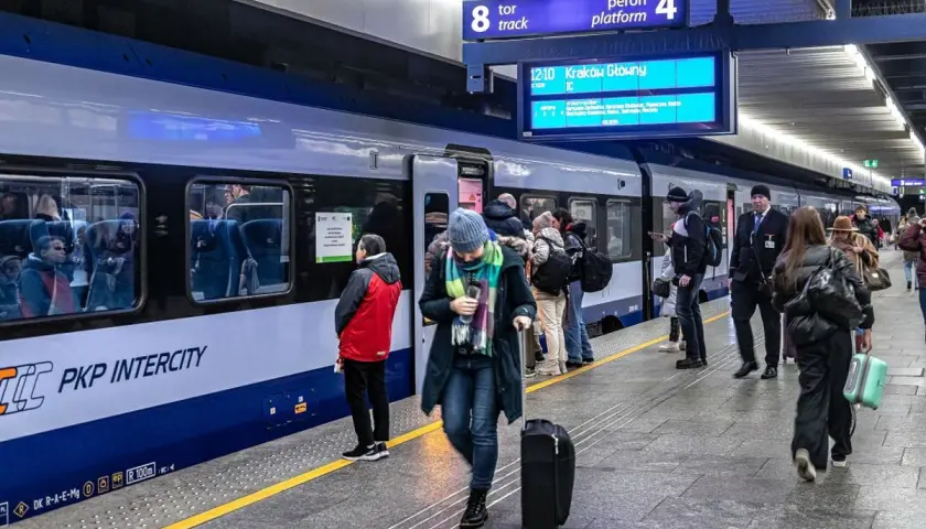 Ludzie z bagażami czekają na pociąg PKP Intercity. Na tablicy informacyjnej widnieje kierunek Kraków Główny. Może to być początek podróży z biletem kredytowym.
