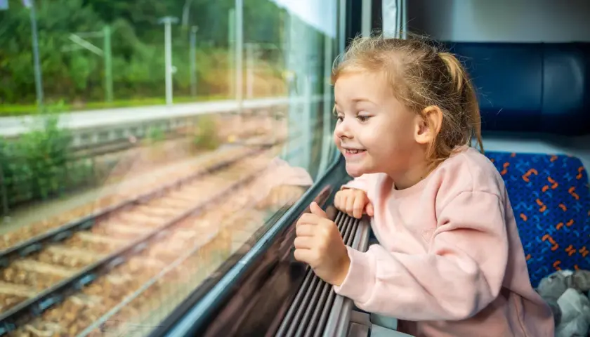 Uśmiechnięta dziewczynka patrzy przez okno pociągu. Zastanawia się, od ilu lat można jeździć samemu pociągiem, marząc o podróżach.