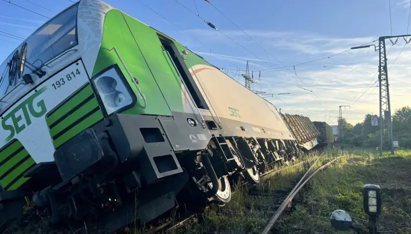 Zielony pociąg towarowy wykoleił się, przechylając się na bok na torach. W tle błękitne niebo i linie energetyczne.