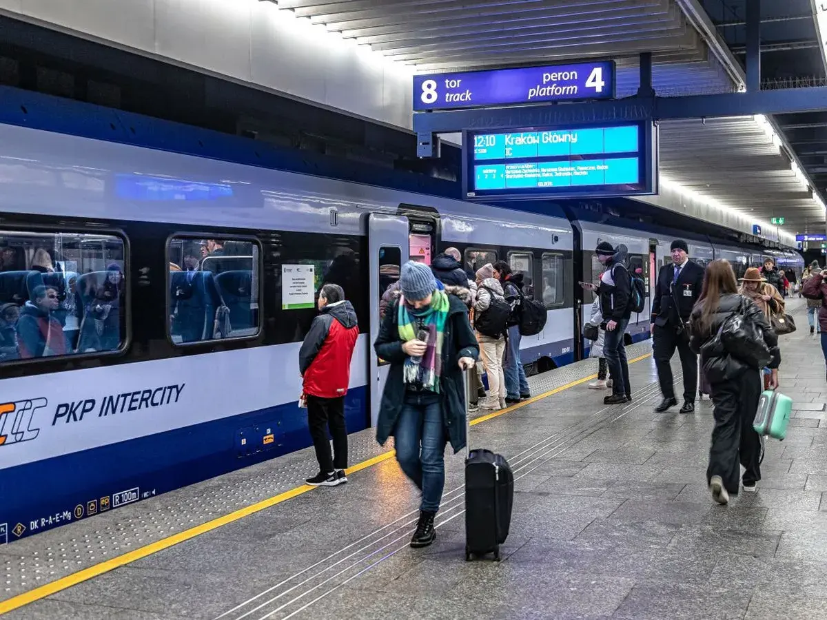 Ludzie z bagażami czekają na pociąg PKP Intercity. Na tablicy informacyjnej widnieje kierunek Kraków Główny. Może to być początek podróży z biletem kredytowym.