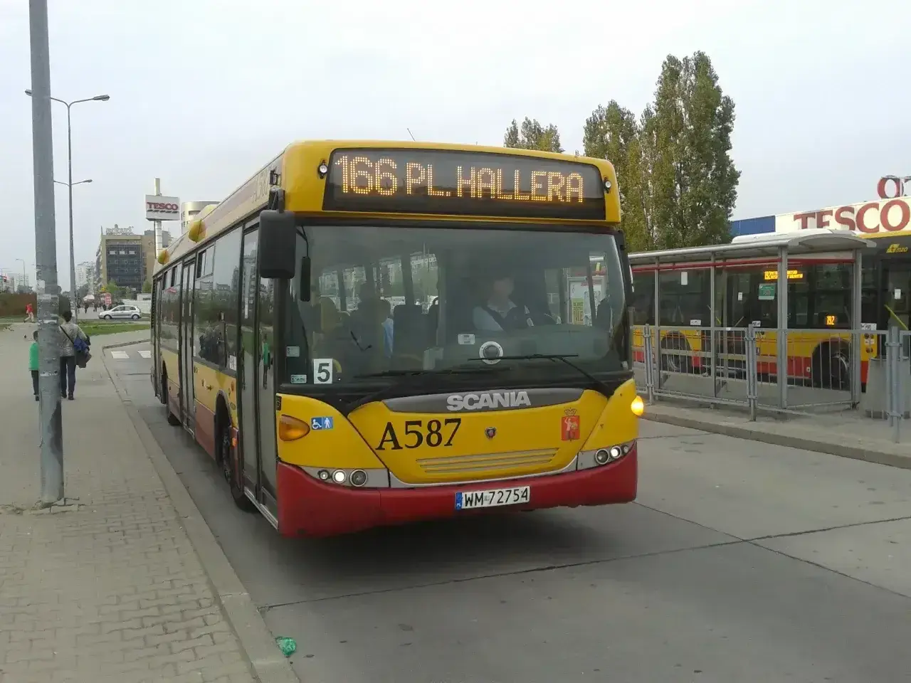 Żółty autobus linii 166 w Warszawie, z napisem "166 PL. HALLERA" na wyświetlaczu, zbliża się do przystanku.